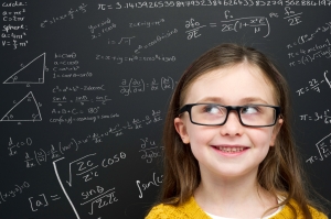 Smart young girl wearing a yellow jumper and glasses stood infront of a blackboard with mathematical equations written in chalk